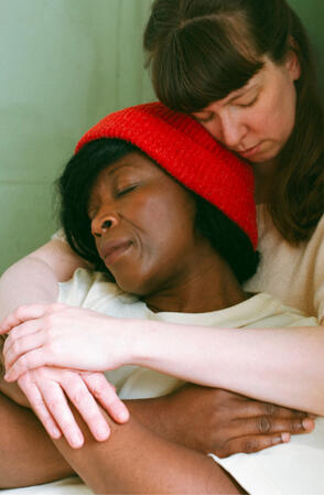 Two women, rest against each other with their arms wrapped softly, over their chests. Tekie Quaye Studio